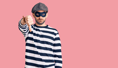 Sticker - Young handsome man wearing burglar mask looking unhappy and angry showing rejection and negative with thumbs down gesture. bad expression.