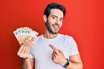 Young hispanic man holding euro banknotes smiling happy pointing with hand and finger