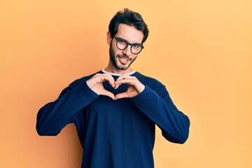 Wall Mural - Young hispanic man wearing casual clothes and glasses smiling in love showing heart symbol and shape with hands. romantic concept.