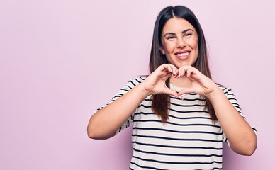 Poster - Young beautiful brunette woman wearing casual striped t-shirt over isolated pink background smiling in love doing heart symbol shape with hands. Romantic concept.