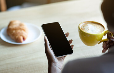 Poster - Unrecognizable black woman holding mobile phone with blank screen while drinking coffee at cafe, mockup for design