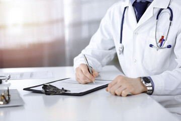 Unknown male doctor sitting and working with clipboard of medication history record in a darkened clinic, glare of light on the background, close-up of hands