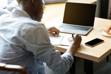 Unrecognizable black guy using laptop with blank screen, studying online, taking notes at cafe, mockup for design