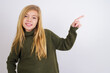 © Roquillo - Caucasian kid girl wearing green knitted sweater against white wall looking at camera indicating finger empty space sales
