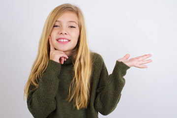 Wall Mural - Funny Caucasian kid girl wearing green knitted sweater against white wall hold open palm new product great proposition