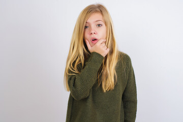 Wall Mural - Caucasian kid girl wearing green knitted sweater against white wall Looking fascinated with disbelief, surprise and amazed expression with hands on chin