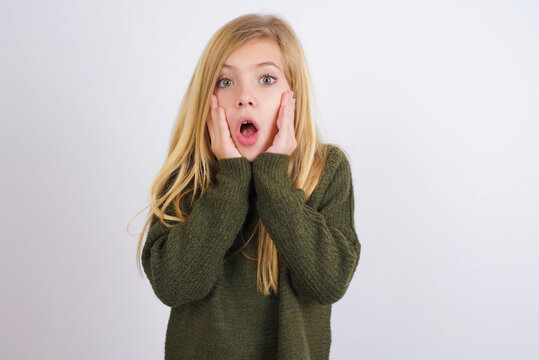 Scared terrified Caucasian kid girl wearing green knitted sweater against white wall  shocked with prices at shop, People and human emotions concept