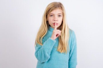 Wall Mural - Cute Caucasian kid girl wearing blue knitted sweater against white wall makes hush gesture, asks be quiet. Don't tell my secret or not speak too loud, please!