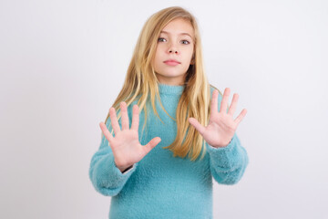 Wall Mural - Cute Caucasian kid girl wearing blue knitted sweater against white wall doing stop sing with palm of the hand. Warning expression with negative and serious gesture on the face.