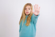 © Jihan - Cute Caucasian kid girl wearing blue knitted sweater against white wall doing stop gesture with palm of the hand. Warning expression with negative and serious gesture on the face.
