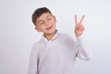 Wall Mural - Cute Caucasian kid boy wearing knitted sweater against white wall smiling with happy face winking at the camera doing victory sign. Number two.