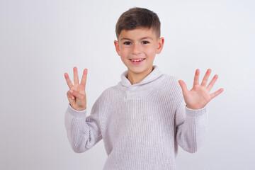 Wall Mural - Cute Caucasian kid boy wearing knitted sweater against white wall showing and pointing up with fingers number eight while smiling confident and happy.