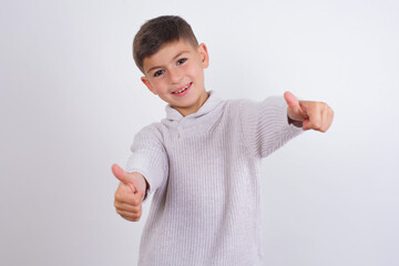 Wall Mural - Cute Caucasian kid boy wearing knitted sweater against white wall approving doing positive gesture with hand, thumbs up smiling and happy for success. Winner gesture.