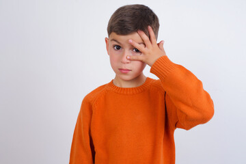 Wall Mural - Cute Caucasian kid boy wearing knitted sweater against white wall peeking in shock covering face and eyes with hand, looking through fingers with embarrassed expression.