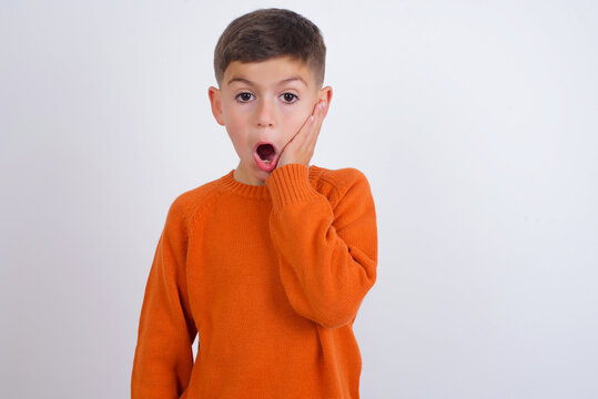 Shocked Cute Caucasian kid boy wearing knitted sweater against white wall looks with great surprisment being very stunned, astonished with unexpected news, Facial expressions concept.