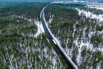 Wall Mural - white truck driving on asphalt road through the winter forest. seen from the air. Aerial view landscape. drone photography.  cargo delivery in winter