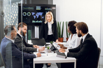 Likable concentrated blond female boss leader at a meeting together with her diverse multiracial colleagues, in modern boardroom with big TV plasma screen, presenting world map and charts