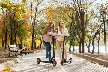 Two young beautiful girls ride electric scooters in the Park on a warm autumn day