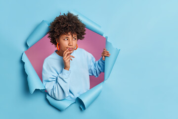 Poster - Photo of thoughtful dark skinned Afro American woman looks away and considers something keeps hand near mouth breaks through paper hole thinks about decision wears casual jumper and earrings