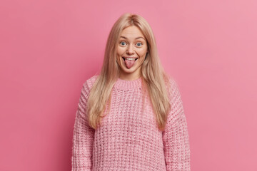 Wall Mural - Funny blonde woman sticks out tongue and makes grimace wears warm knitted sweater poses against pink background has fun mimicks childish faces stays happy and positive. Human face expressions