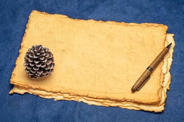 Wall Mural - stack of tan toned heavyweight deckle edge paper sheets with a stylish pen and decorative frosty pine cone - holiday greeting card