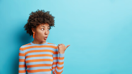 Wall Mural - Studio shot of surprised curly haired woman keeps mouth opened points thumb away on blank space wears striped jumper shows amazing offer isolated over blue background. Advertisement concept.