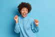 © wayhome.studio  - Photo of joyful dark skinned woman dances carefree keeps fists raised looks positively aside dressed in casual jumper moves against blue background. People positive emotions and feelings concept