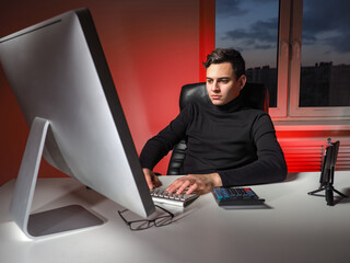 Poster - Remote work of a financial analyst. Man works at the computer in the evening. Overtime work. Irregular working day of a young businessman. Person works at a computer in red light. Fatigue from work.