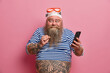 © wayhome.studio  - Positive blue eyed man has long thick beard raises fist holds modern cellphone wears swim goggles and striped sailor jumper has big tattooed belly isolated over pink background. Summer holidays
