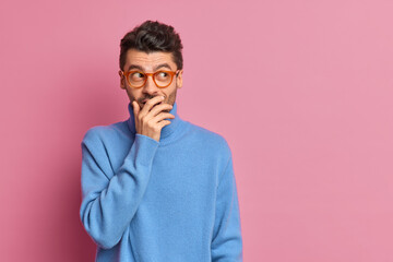 Wall Mural - Studio shot of happy man tries to hide emotions giggles silently and covers mouth with hand looks aside positively dressed in casual clothes poses against pink background blank copy space on right