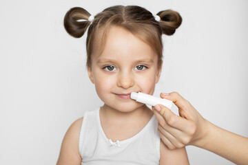 Lips hygiene and care. Sunscreen, wind and frost protection of kids lips. Close up horizontal studio shot of mom's hand, applying lip balm on the lips of happy smiling pretty girl, isolated on white