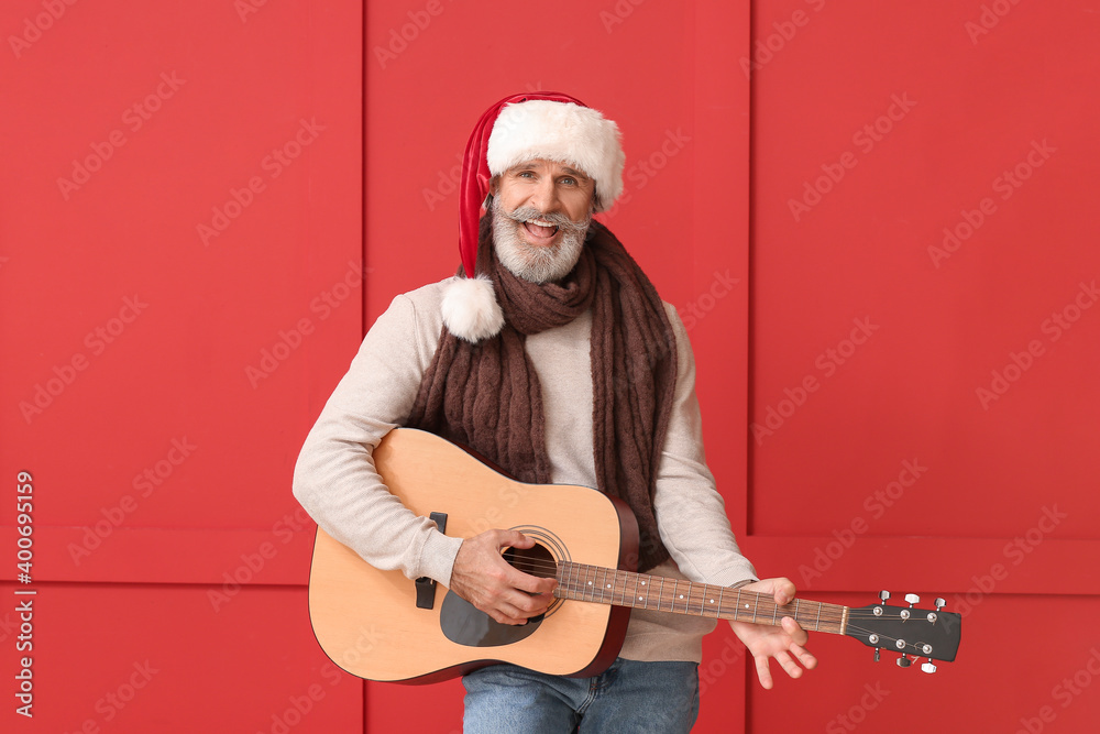 Senior man in Santa hat playing guitar on color background