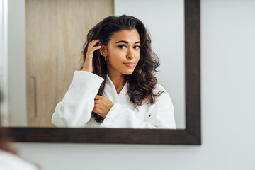 Wall Mural - Beautiful female wearing bathrobe looking at a mirror and touching her hair