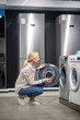 © zinkevych - Woman in striped shirt choosing a washing machine in a showroom and looking interested