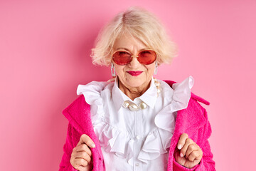 fancy aged lady in eyeglasses posing at camera wearing fashionable clothes, isolated over pink backg