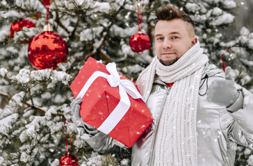 Young bearded man in down coat stands near decorated Christmas New Year tree fir holding holiday gift box gesturing like sign outdoors in winter snowy forest and nature at background. New Year mood