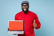© ViDi Studio - Smiling young bearded african american man 20s in casual red shirt eyeglasses cap showing thumb up hold laptop pc computer with blank empty screen isolated on pastel blue background studio portrait.