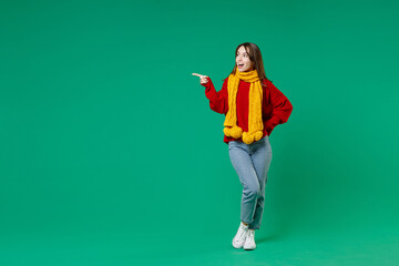 Wall Mural - Full length of funny young brunette woman 20s wearing casual knitted red sweater yellow scarf standing pointing index finger aside looking camera isolated on green color background studio portrait.