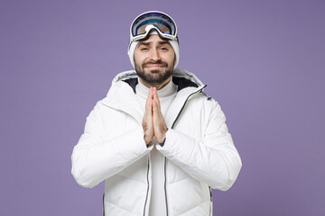 Wall Mural - Pleading skier man in white windbreaker jacket ski goggles mask hold hands folded in prayer spend extreme weekend winter in mountains isolated on purple background. People lifestyle hobby concept.