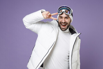 Wall Mural - Cheerful skier man in white windbreaker jacket ski goggles mask showing victory sign spend extreme weekend winter season in mountains isolated on purple background. People lifestyle hobby concept.