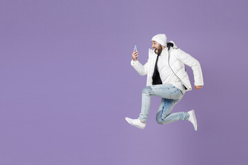 Wall Mural - Full length side view shocked man in warm white padded windbreaker jacket hat jump using mobile cell phone isolated on purple background studio portrait. People lifestyle cold winter season concept.