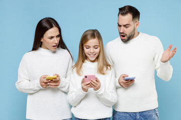 Wall Mural - Shocked young parents mom dad with child kid daughter teen girl in white sweaters using mobile phone typing sms message isolated on blue background studio. Family day parenthood childhood concept.