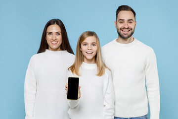 Wall Mural - Smiling young parents mom dad with child kid daughter teen girl in white sweaters hold mobile phone with blank empty screen isolated on blue background studio. Family day parenthood childhood concept.