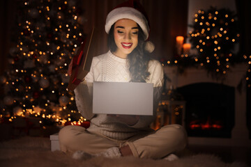 Canvas Print - Young woman wearing Santa hat opening Christmas gift on floor at home