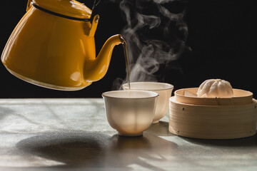 Steam from two cups and yellow pot has a fragrant tea and steamed buns in wooden basket, Morning tea on black background,Food that provides warmth to body and has health benefits in the winter season.