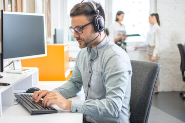 Wall Mural - Handsome male customer support phone operator with headset working on desktop computer in office.