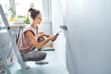 Young Woman Painting Wall With Paint Roller While Crouching At Home