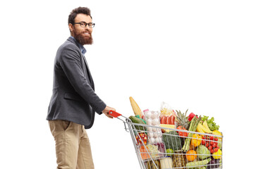 Wall Mural - Young bearded man with glasses pushing a shopping cart with food products and smiling at the camera