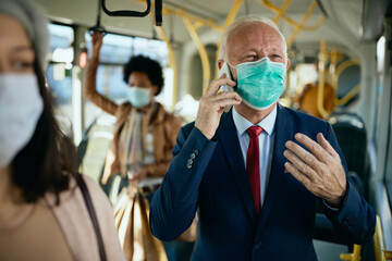 Wall Mural - Happy senior businessman with face mask talking on the phone in a public transport.