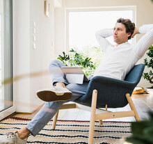 Mid Adult Man With Hands Behind Head Sitting On Chair At Home